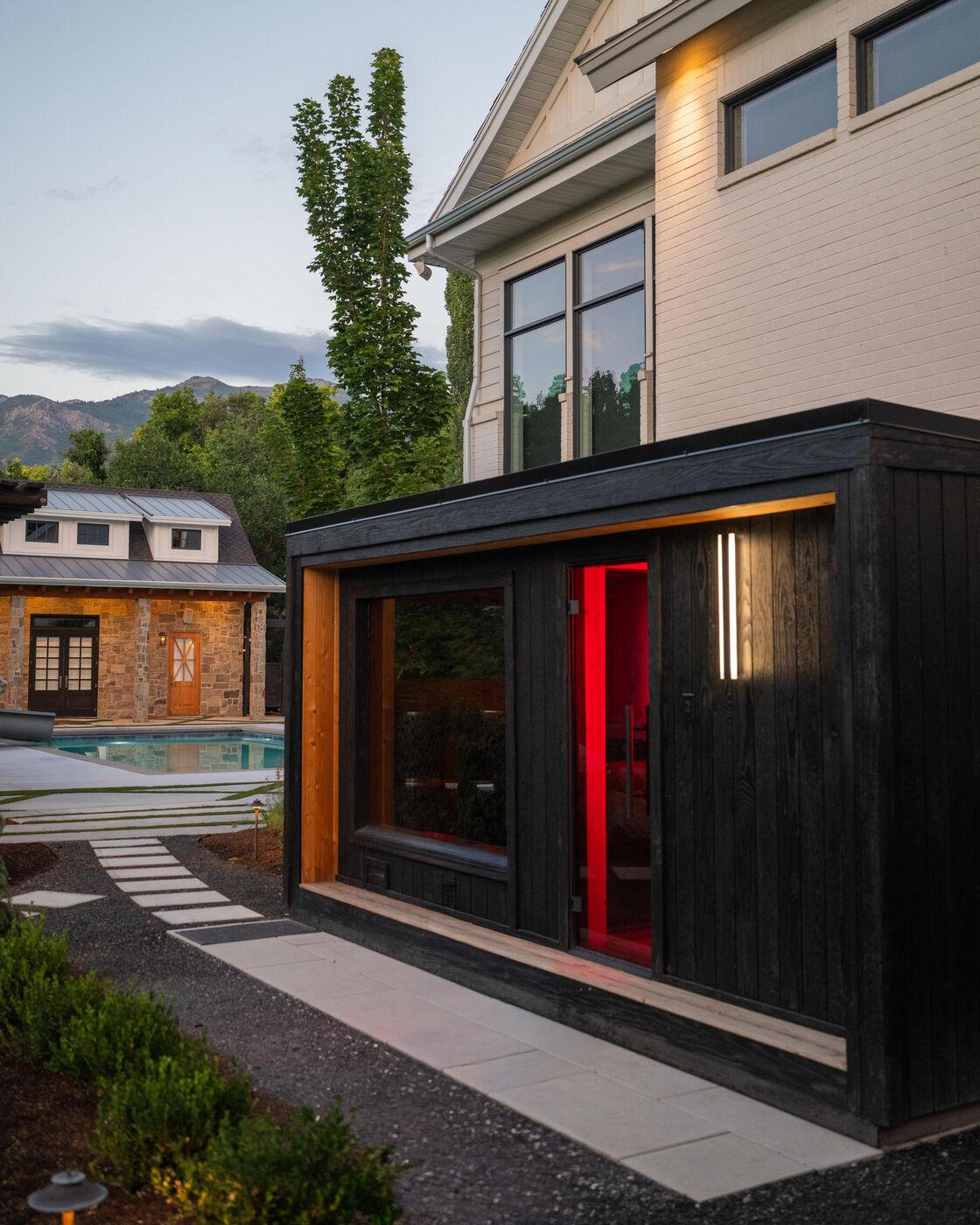 A blackened-timber studio lit from within at dusk, the pool visible beyond it
