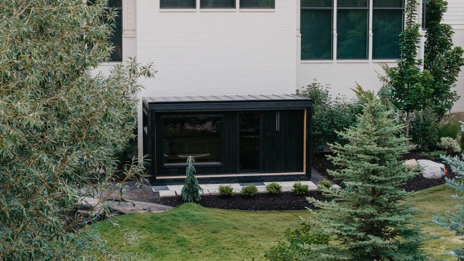 A blackened structure framed by evergreens and mulch beds, with clipped planting meeting a sod lawn