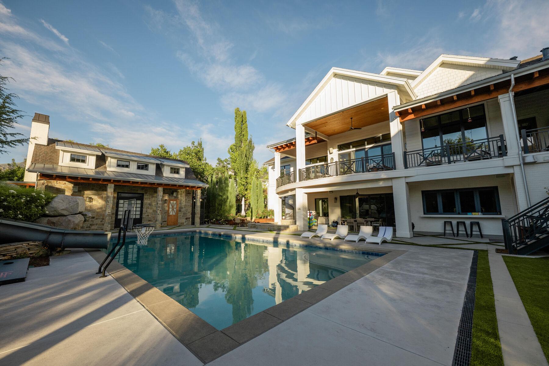 A rectangular pool running the length of a stone-and-siding house, with a mown lawn strip and the mountains beyond