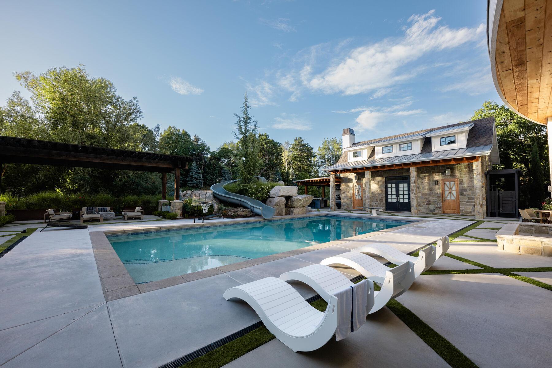 Curved loungers on a broad pool deck beside a stone pool house