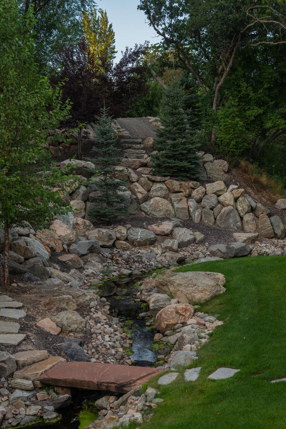 A stone-edged stream crossed by a timber bridge, running out onto mown lawn